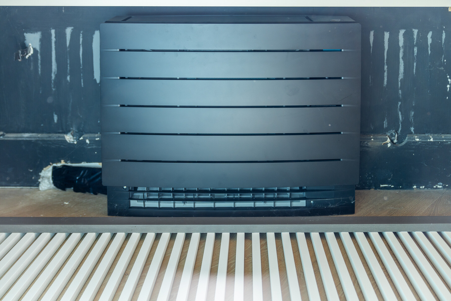 Detail van de matzwarte cover van een Daikin Perfera-vloermodel achter een lattenrooster in Brugge